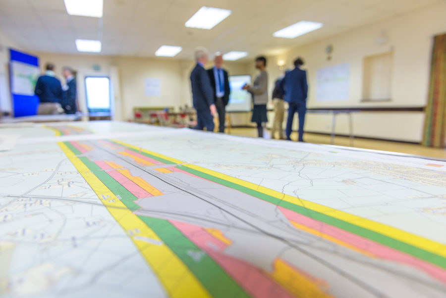 a long map on the table in the foreground with a group of standing people behind talking.